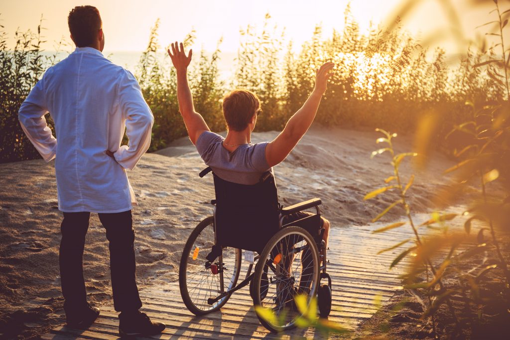 A man on wheelchair and his nurse enjoying sunrise on a beach.